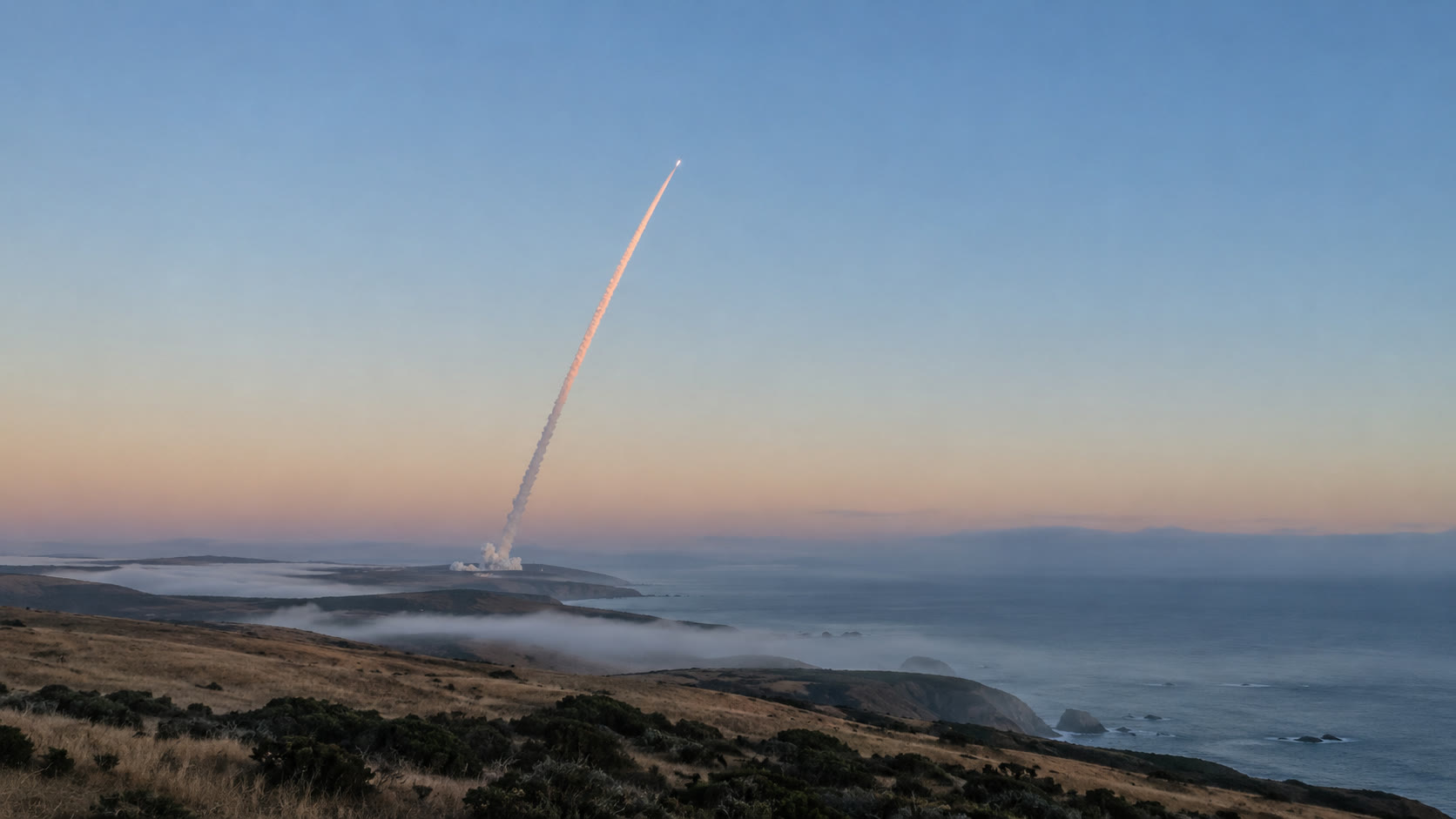 A rocket's exhaust trail curving up into a pale dawn sky, seen from far away across still water.