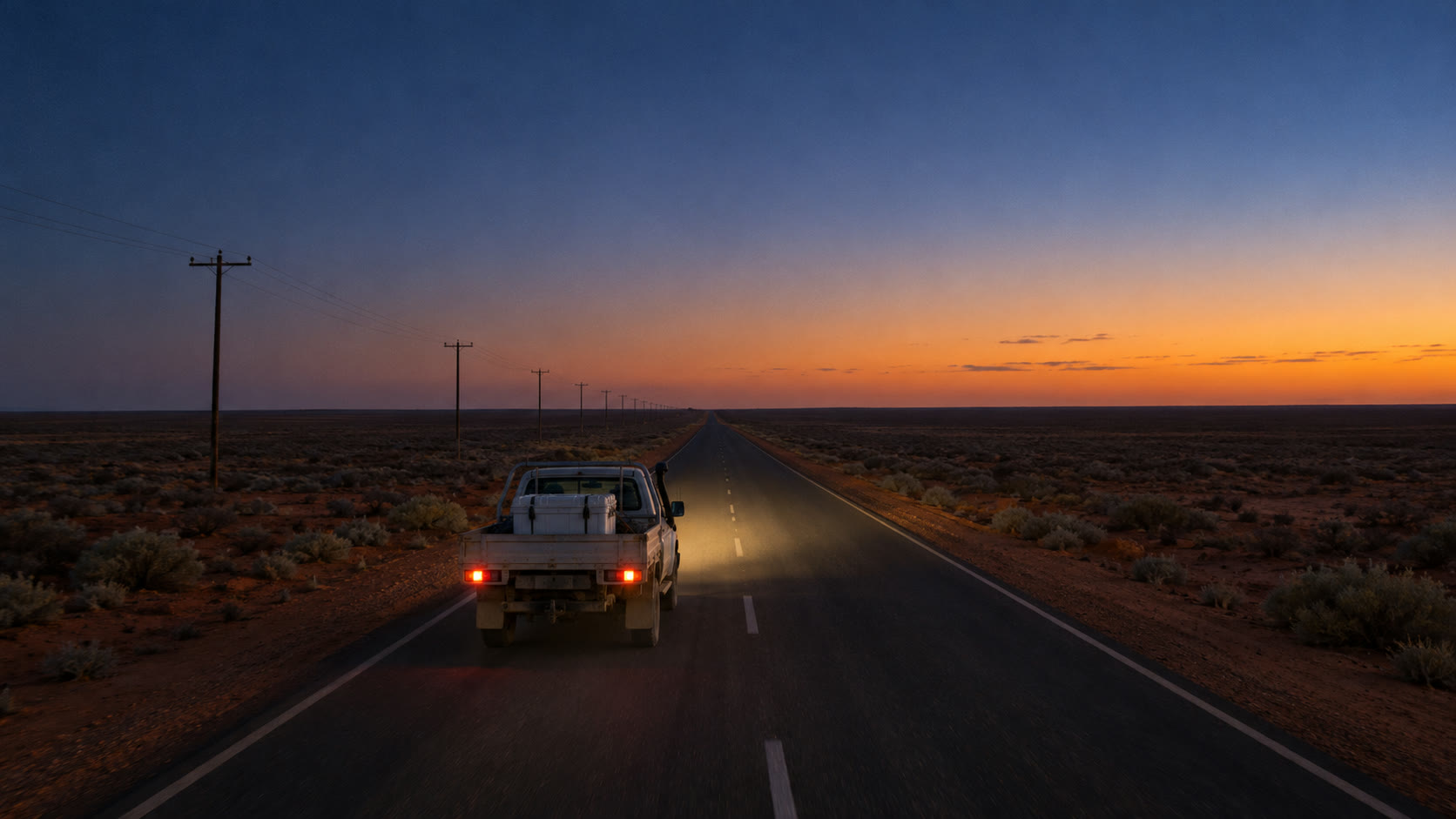 A white ute drives a long straight outback highway at dusk.
